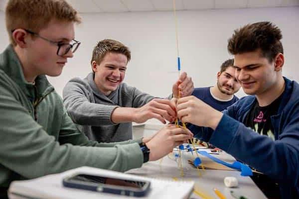 A group of four male students work together to construct a tower out of raw spaghetti and blue painter’s tape in a classroom.